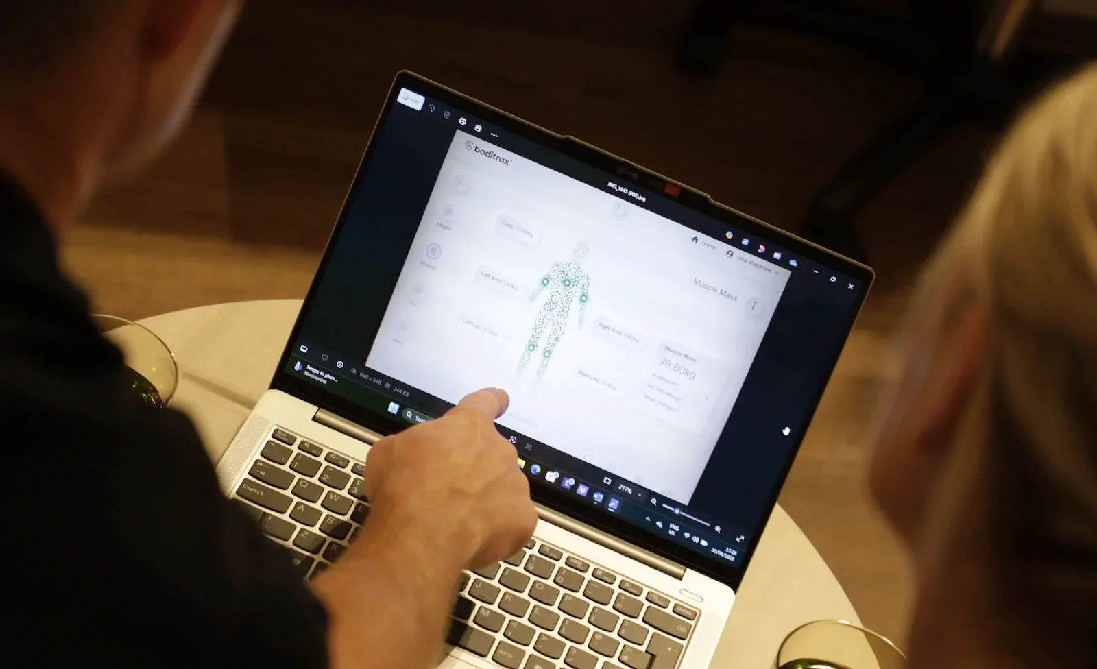 A person pointing at a laptop screen that displays a human body diagram with green dots or data points mapped across it. The laptop appears to be sitting on a light surface, and someone in dark clothing is interacting with what looks like a health or medical interface.